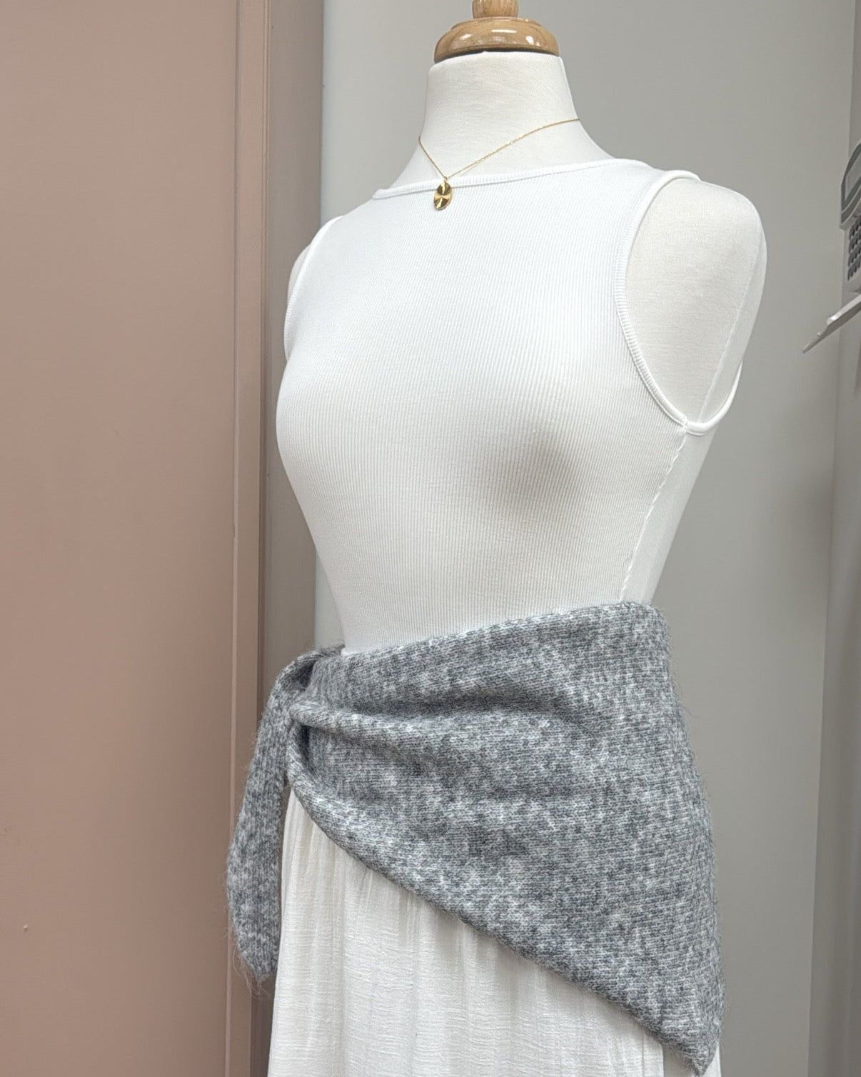 A gray knitted wrap or sash tied around the waist of a white sleeveless dress displayed on a mannequin torso, photographed indoors against a plain wall.